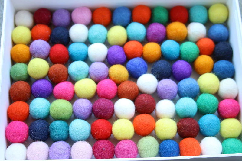 A white rectangular box filled with many small felt pom-poms in assorted bright colors, including red, orange, yellow, green, blue, purple, pink, and white.