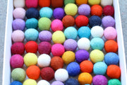 A close-up view of a white tray filled with many small felt pom-poms in assorted bright colors, including pink, blue, yellow, orange, red, purple, green, and white.