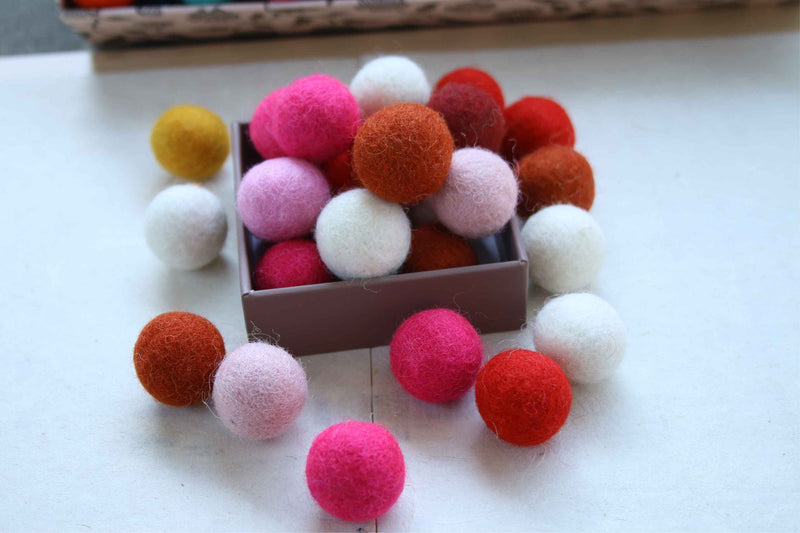 A collection of colorful felt pom-poms in shades of pink, red, orange, white, and yellow, with some scattered around and others piled inside a small brown box on a white surface.