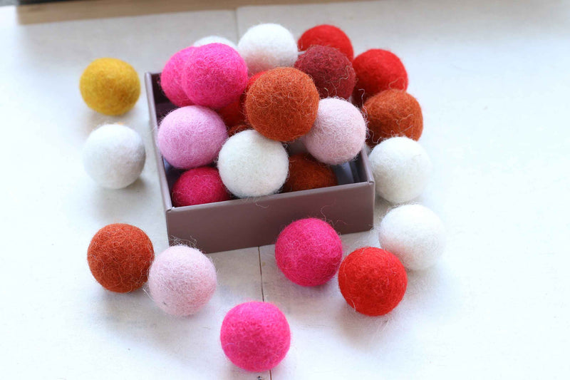 A collection of colorful wool felt pom-poms in shades of pink, red, orange, white, and yellow, with some scattered around and others piled inside a small brown box on a white surface.