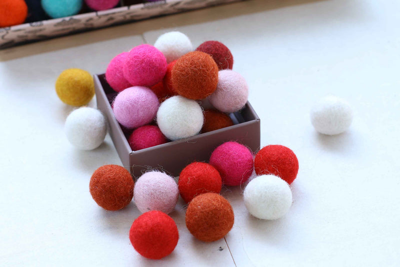 A small box filled with colorful felt pom-poms in red, orange, pink, white, and yellow, with some scattered around on a white surface.