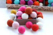 A collection of colorful felt pom-poms in various shades of pink, orange, red, white, yellow, and purple, displayed in a small box with more of the same in the background.