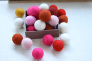 A collection of colorful felt pom-poms in pink, red, orange, white, and yellow, with some scattered around a small box on a white surface.