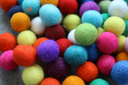 A close-up pile of small felt pom-poms in assorted bright colors including orange, pink, blue, green, purple, yellow, and red on a gray surface.