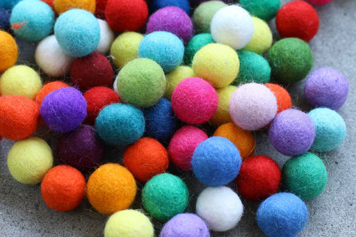 A pile of colorful felt pom-poms in various shades including blue, green, yellow, orange, pink, purple, and white, resting on a gray textured surface.