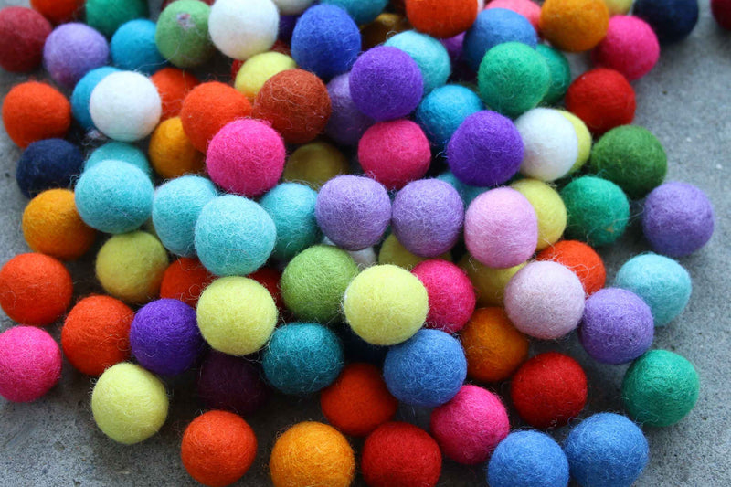 A pile of colorful felt pom-poms in various colors including pink, blue, green, yellow, orange, purple, and white, arranged on a gray textured surface.