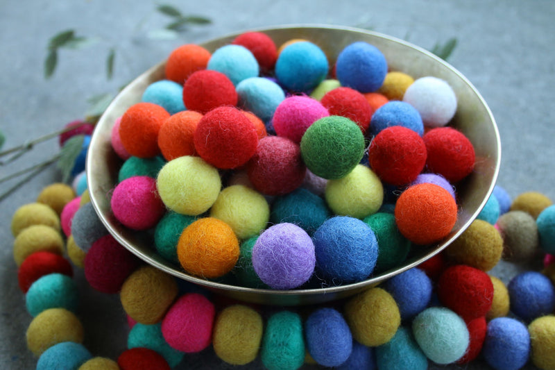 A bowl filled with colorful felt pom-poms in assorted shades including red, blue, green, yellow, pink, orange, and purple, arranged on a gray surface.