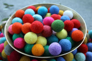 A metal bowl filled with colorful felt pom-poms in red, orange, blue, green, pink, yellow, purple, and white, set against a soft gray background.