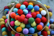 A metal bowl filled with colorful felt pom-poms in red, orange, yellow, green, blue, pink, purple, and white, with more pom-poms scattered around on a gray surface.