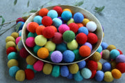 A bowl filled with colorful felt pom-poms, with additional multicolored pom-poms arranged around it on a gray surface.