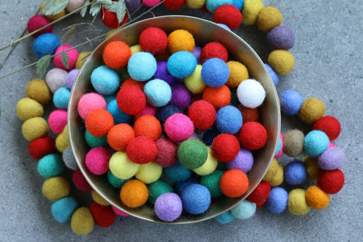 A metal bowl filled with colorful felt pom-poms, with more of the same scattered around it on a gray surface.
