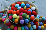A metal bowl filled with colorful felt pom-poms, with more of the same scattered around it on a gray surface.