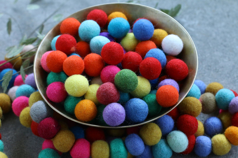 A metal bowl filled with colorful felt pom-poms, with more of the same scattered around it on a gray surface.