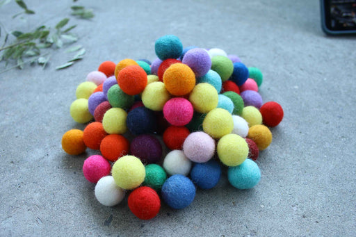A pile of colorful felt pom-poms in various colors including orange, yellow, green, blue, pink, purple, and white, arranged on a gray concrete surface.