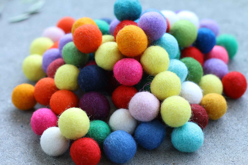 A pile of small, colorful felt pom-poms in various shades including orange, yellow, green, blue, pink, purple, and white, resting on a textured gray surface.