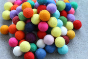 A pile of colorful felt pom-poms in various shades including orange, yellow, green, blue, pink, purple, and white, resting on a gray textured surface.