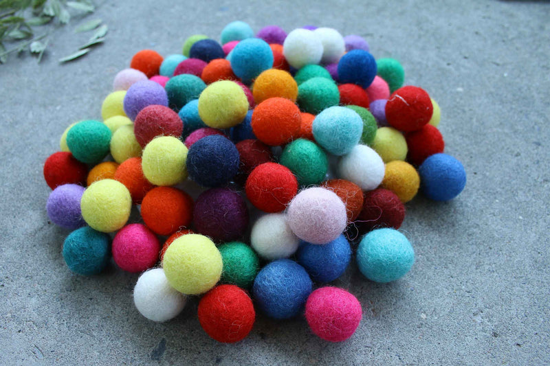 A pile of colorful felt pom-poms in various colors including red, blue, green, yellow, pink, orange, purple, and white, arranged on a gray concrete surface.
